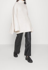 Overmaatse geribbelde gebreide coltrui in crème, gecombineerd met zwarte faux leren wijde broek en chunky zwarte loafers.