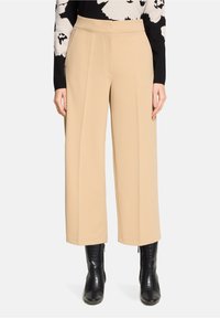Pantalon beige à jambes larges avec une texture lisse, taille haute et plis sur le devant, associé à des bottines noires embossées façon crocodile.