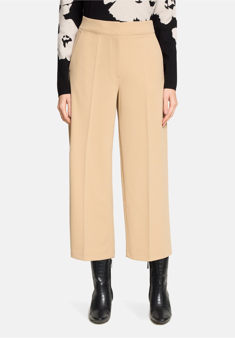 Pantalon beige à jambes larges avec une texture lisse, taille haute et plis sur le devant, associé à des bottines noires embossées façon crocodile.