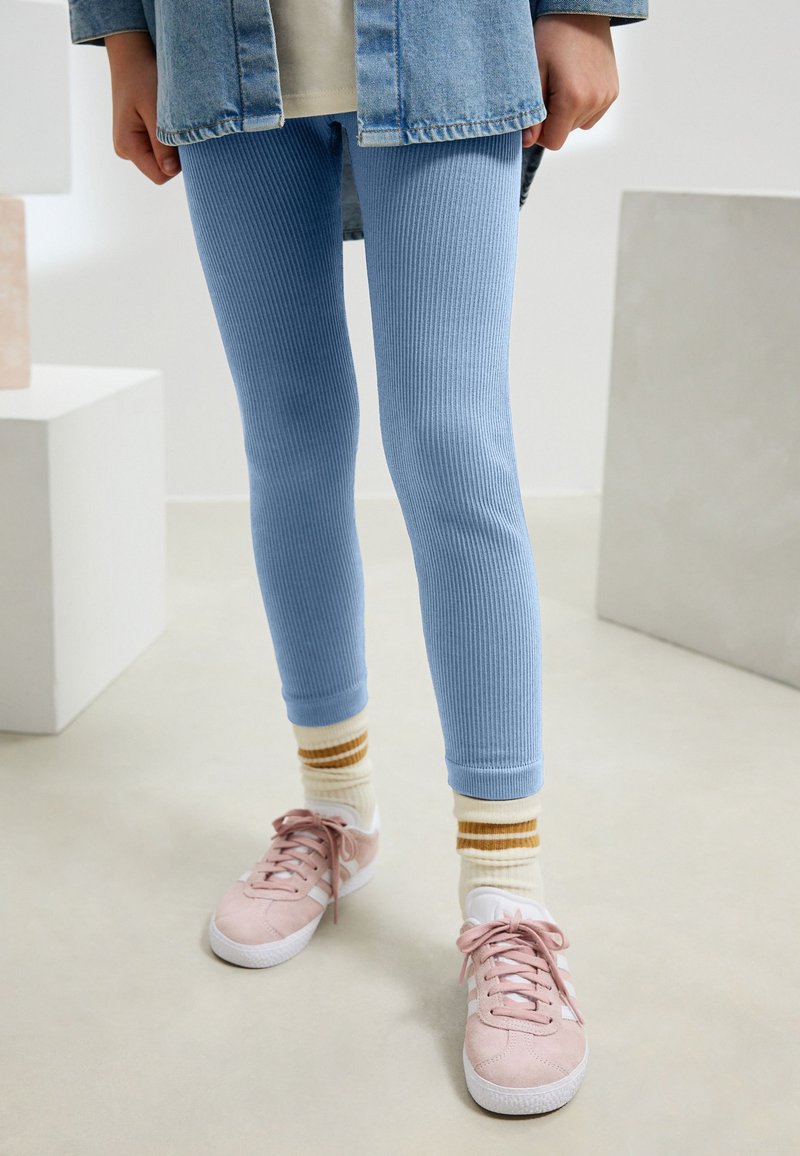 Hellblaue, gerippte Leggings mit eng anliegendem Schnitt und kontrastierenden hellblauen Bündchen. Kombiniert mit pinken Turnschuhen und gestreiften Socken.