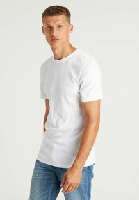 Biała koszulka z okrągłym dekoltem z bawełnianej tkaniny, z krótkimi rękawami, z subtelnym detalem logo na klatce piersiowej. Noszona z jasnoniebieskimi dżinsami.