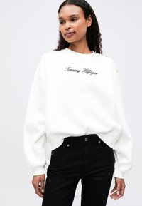 Valkoinen collegepaita, jossa on brodeerattu "Tommy Hilfiger" -logo, kultaiset nappikoristeet olkapäillä ja resorit hihoissa. Yhdistetty mustiin housuihin.