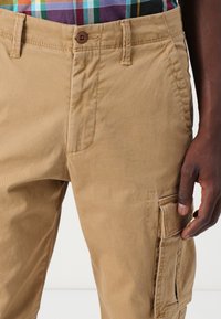 Khaki cargo pants featuring a button closure, side pockets with flaps, and a textured finish. The waistband is stitched for durability.