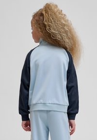 Laps, kellel on pikad lokkis juuksed, kannab helesinist ja musta spordijopet ning sobivaid helesiniseid pükse, seisab seljaga ühtlase tausta ees.