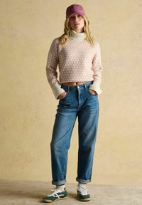 Frau mit einem rosa gemusterten Rollkragenpullover, blauen hochgekrempelten Jeans, grün-weißen Turnschuhen und einer rosa Kappe, die mit den Händen in den Taschen dasteht.