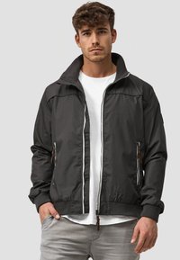 Black bomber jacket with a zip front, ribbed cuffs, and hem. Features two side pockets with brown leather pulls. White t-shirt underneath.