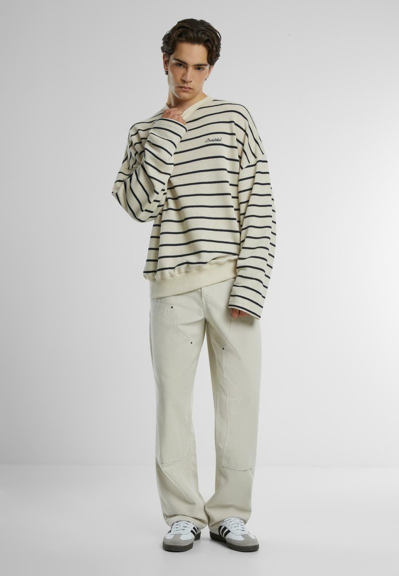 Felpa oversize a righe crema e blu navy con polsini e orlo a coste, abbinata a pantaloni beige chiaro. Calzature casual completano il look.