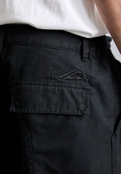 Black cargo pants with flap pocket and embroidered brand logo worn with white shirt, close-up on hip and waist area.