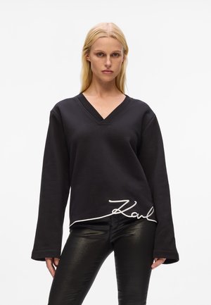 Felpa nera oversize con scollo a V, maniche lunghe e firma "Karl" ricamata in bianco sull'orlo; texture liscia.