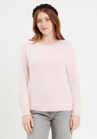 Lichtroze, fluffy trui met lange mouwen en een ronde hals. Zachte textuur en casual pasvorm, gecombineerd met een zwarte haarband.
