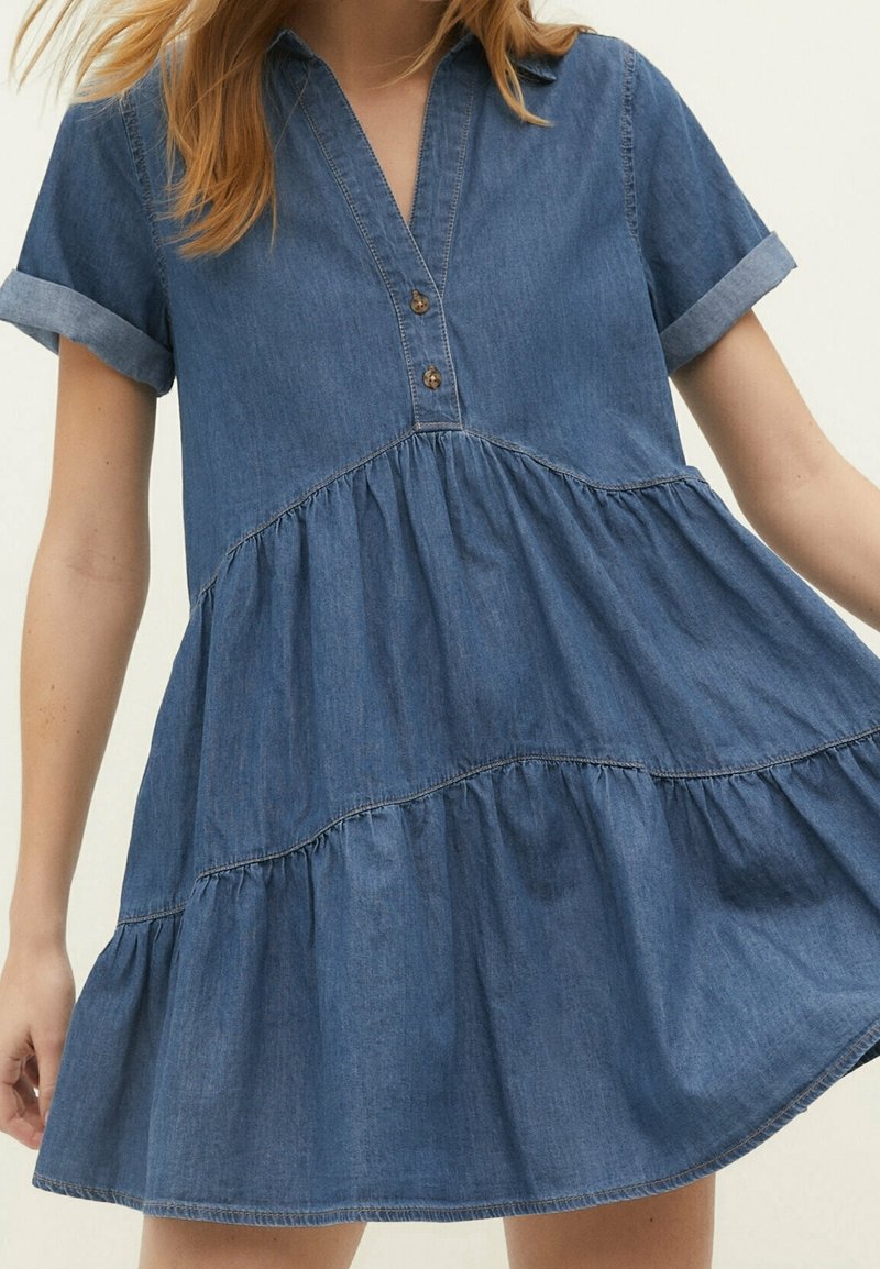 Femme portant une robe en jean bleu à manches courtes avec une jupe à volants, un col en V boutonné et des manches retroussées, montrée des épaules jusqu'à mi-cuisse.