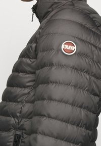 Grå puffjacka med horisontellt quiltat mönster, dragkedjestängning och en rund "Colmar" logotyp patch på övre vänstra sidan.