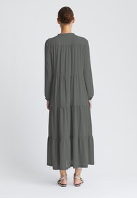 Robe longue grise à volants avec manches longues, taille froncée et ourlet arrivant aux chevilles. Texture lisse et design simple. Sandales portées en dessous.