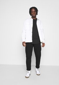 Jachetă albă cu nasturi peste un tricou negru, pantaloni negri și adidași albi cu accente maro. Modelul stă pe un fundal simplu.
