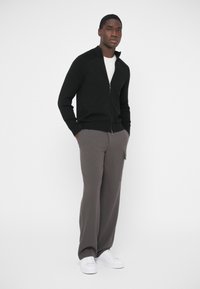 Čierny kardigán na zips s rebrovanými akcentmi na pleciach, v kombinácii so sivými voľnými nohavicami a bielymi teniskami. Jednoduchý, moderný dizajn.