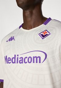 Weißes Sporttrikot mit einem lila Kragen und Akzenten, strukturiertem Muster, lila 'Mediacom'-Logo und lila Vereinswappen.