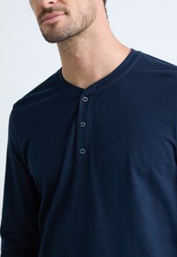 Man in een donkerblauw lange mouwen henley shirt met drie knopen, met een zichtbare nek en gedeeltelijk ondergezicht tegen een effen achtergrond.