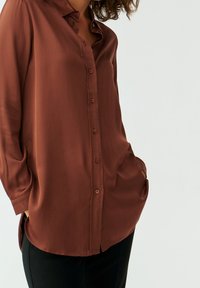 Rust-colored blouse made of smooth fabric; features a collar, button front, and loose fit with a curved hem. Black pants visible at bottom.