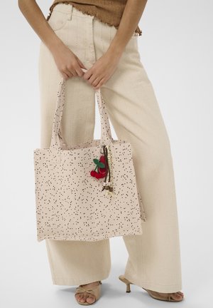Femme tenant un sac fourre-tout beige avec des taches noires et un porte-clés cerise, portant un pantalon large crème et des talons ouverts beige.
