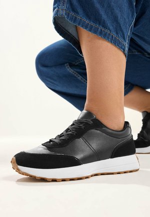 Zapatilla negra y gris con suela blanca y banda de goma, usada con vaqueros azules remangados, pie apoyado en el suelo contra un fondo blanco roto.