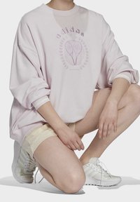 Ljusrosa oversized sweatshirt med lila grafiskt tryck. Ribbad halsringning och muddar. Bärs med beige shorts och vita sneakers.