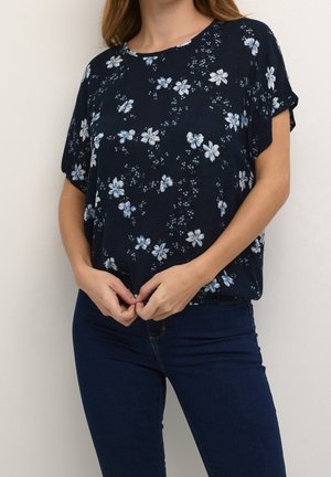 Femme portant un chemisier ample bleu marine à motifs floraux avec des manches courtes et un jean bleu foncé, mains jointes à la taille.