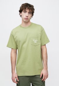 T-shirt en coton vert olive à manches courtes, col rond, avec une poche poitrine ornée d'un logo brodé blanc.