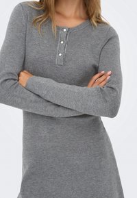 Femme aux cheveux châtain clair porte une robe en maille grise ajustée à manches longues avec une encolure henley à trois boutons, les bras croisés.