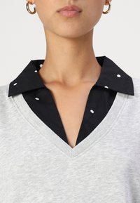 Felpa grigia caratterizzata da un scollo a V, sovrapposta a un colletto nero a pois. Tessuto testurizzato e design a colori contrastanti.