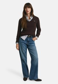 Brauner Kabelstrickpullover, darunter ein hellblaues Hemd mit Kragen, locker sitzende blaue Jeans und dunkle Schuhe. Stehende Pose, Hände in den Taschen.