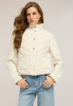 Frau mit langen, welligen Haaren, die eine cremefarbene gesteppte Jacke mit Druckknöpfen, blaue Jeans, ein goldenes Armband und hängende Ohrringe trägt und mit den Händen in den Taschen posiert.