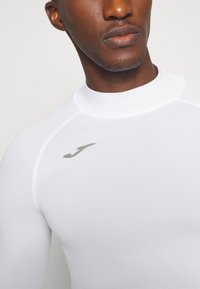White athletic long-sleeve shirt with a high collar and light gray logo on the chest. Smooth texture and form-fitting design.
