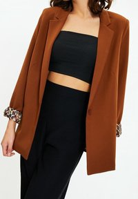 Blazer marrón de manga larga con puños de estampado de leopardo, combinado con una camiseta de corte crop negra de canalé y pantalones negros de cintura alta.