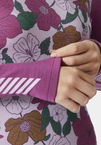 Dlouhý rukáv košile s fialovým květinovým vzorem, texturovanou tkaninou a růžovým manžetovým lemem s bílými pruhy. Detailní záběr ruky na rukávu.