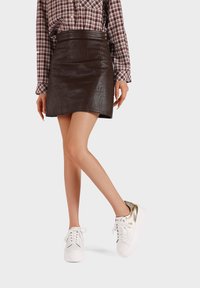 Mini gonna in similpelle marrone abbinata a sneakers bianche con dettagli dorati. La gonna presenta un design aderente e una texture sottile.