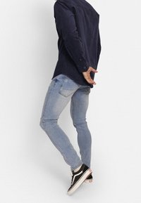 Jeans de mezclilla de color azul claro ajustados con ligeras desgastes, combinados con una camisa de botones azul oscuro y zapatillas negras con suelas blancas.