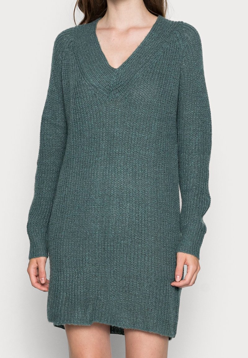 Robe pull en maille couleur bleu sarcelle profond avec col en V, manches longues et texture côtelée, tombant au-dessus du milieu de la cuisse. Coupe décontractée et ample.