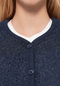 Marineblå teksturert cardigan med rund hals, med to mørke knapper og et kontrasterende hvitt innerlag synlig ved kragen.