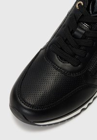 Black athletic shoe with a textured, perforated leather upper, featuring flat black laces, a shiny black lining, and a white strip on the sole.