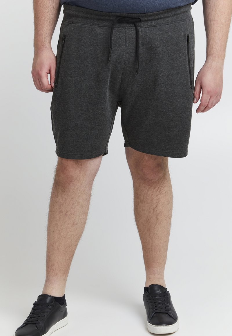Grå shorts laget av mykt stoff, med strikk i livet og to glidelåslommer på sidene. Brukes med svarte joggesko.