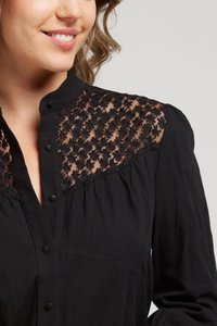 Blouse noire avec empiècement en dentelle florale transparente, col montant et fermeture à boutons. Le tissu est doux avec une finition légèrement texturée.