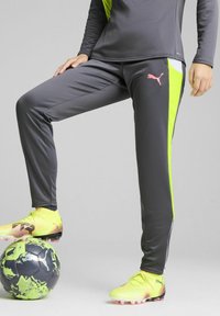 Puma sportska odjeća sadrži tamnosive hlače s neon zelenim bočnim naglascima, u paru s svijetložutim nogometnim kopačkama na crno-zelenoj nogometnoj lopti.