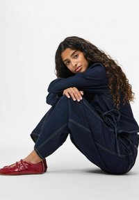 Tumši zila džinsa apģērbs ar jostu viduklī un kontrastējošu dzeltenu šuvju apdari, komplektā ar spīdīgiem sarkaniem baleta apaviem. Seju ietver cirtaini mati.