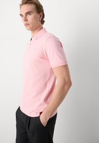 Mladý muž se světlými vlasy, který nosí růžové krátké polo tričko a černé kalhoty, stojí s jednou rukou v kapse na bílém pozadí.