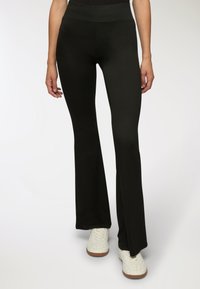 Kobieta w dopasowanych czarnychspodniach z rozszerzanymi nogawkami i białych sznurowanych trampkach, stojąca na tle jasnego, jednolitego tła.