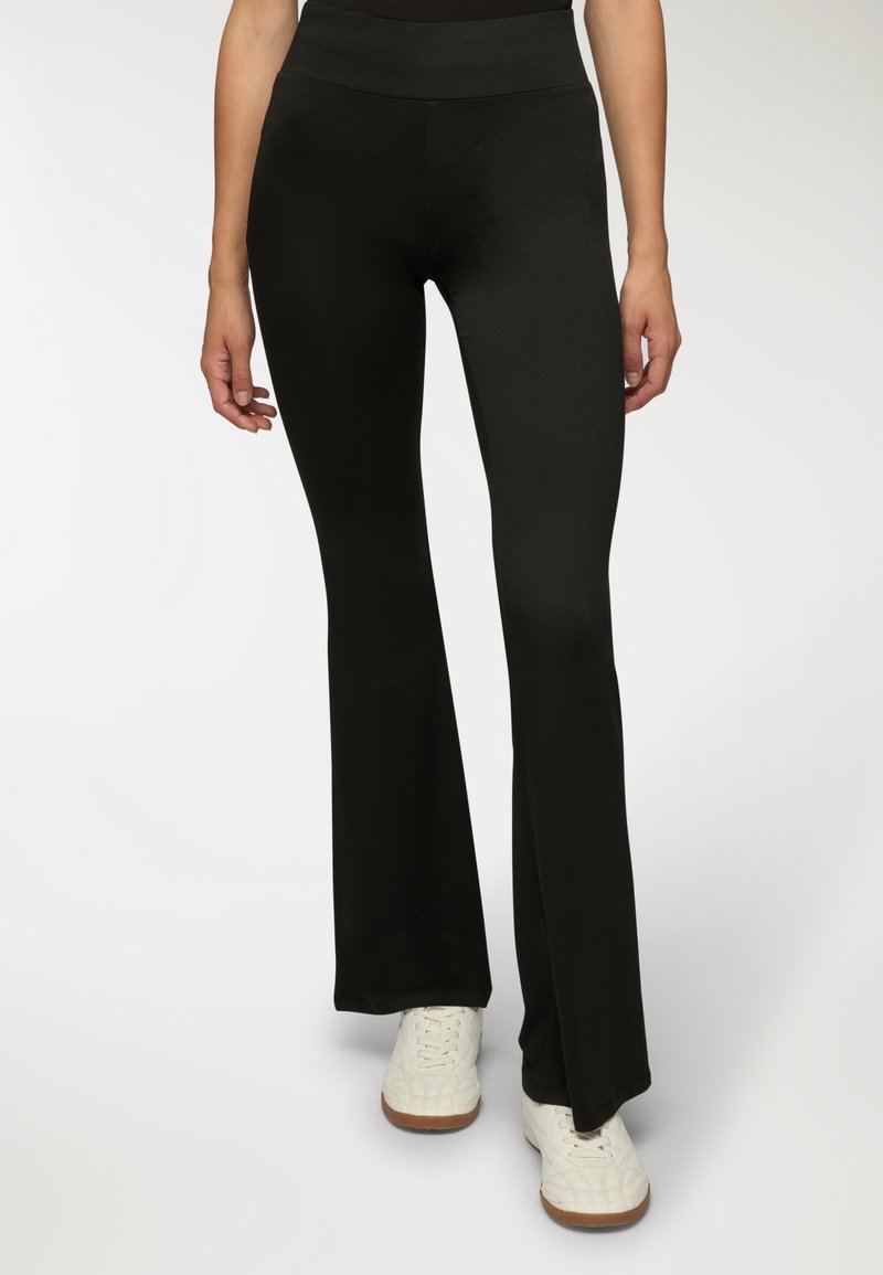 Kobieta w dopasowanych czarnychspodniach z rozszerzanymi nogawkami i białych sznurowanych trampkach, stojąca na tle jasnego, jednolitego tła.