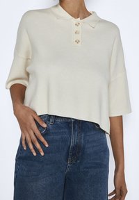 Krémový polo top s krátkým rukávem, límečkem a čtyřmi velkými knoflíky, texturovaná látka, zkrácený střih, spárovaný s modrými džíny.