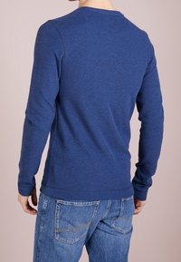 Blå långärmad skjorta med rund halsringning, räfflade detaljer vid axlarna och nederkanten, parat med ljusblå denimjeans.