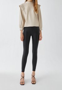 Beige langmouwtop met brede schouderaccenten, gecombineerd met strakke zwarte leggings en zwarte sandalen met hakken en bandjes. De gehele outfit heeft een gladde textuur.