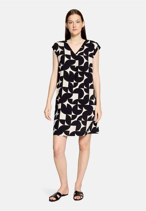Jeune femme debout, portant une robe à manches courtes à motif géométrique noir et blanc et des sandales plates noires, fond neutre.